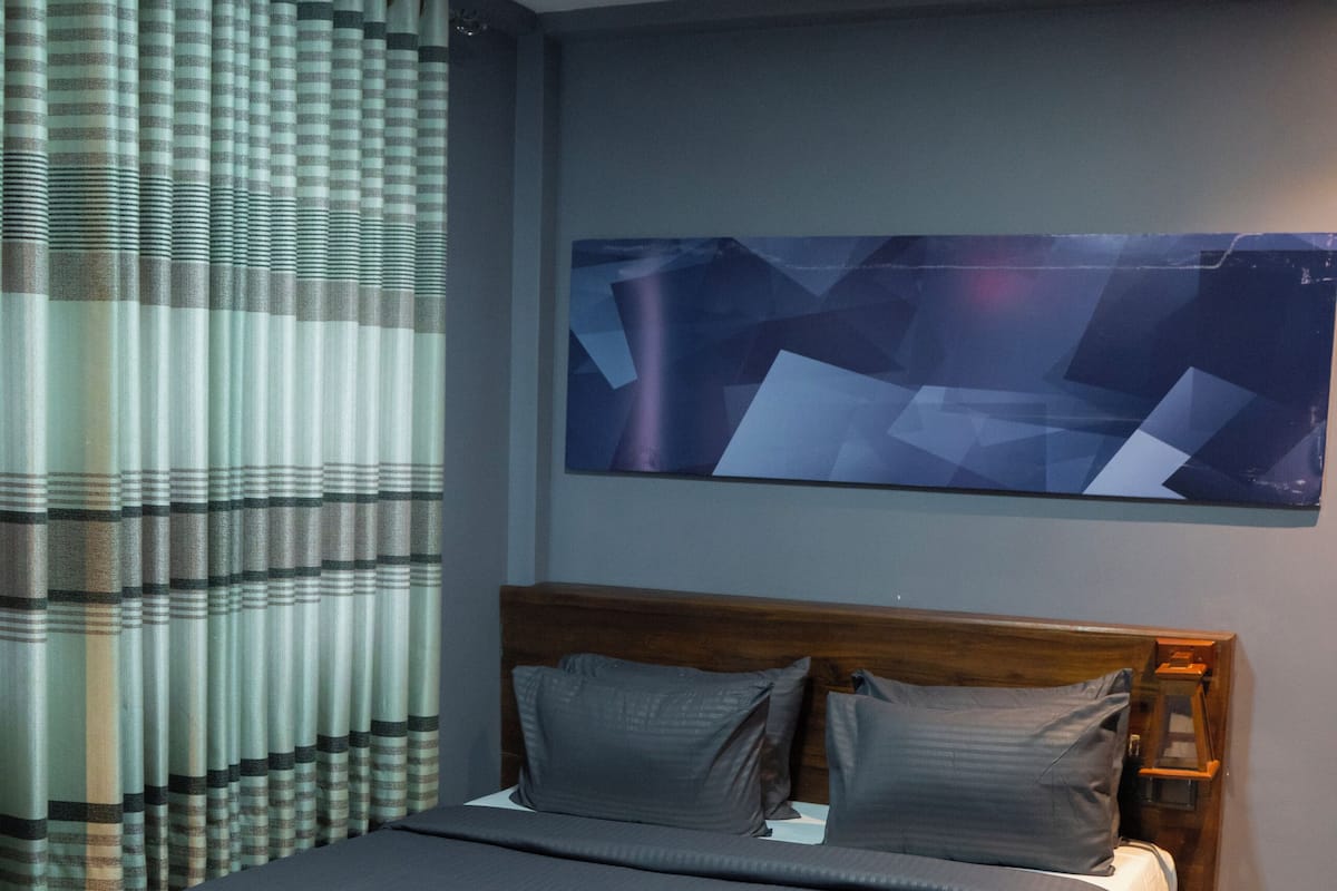 Uma cama bem arrumada com colcha escura e travesseiros, uma cabeceira de madeira e uma obra de arte abstrata pendurada na parede.