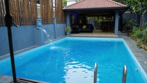 Ein Pool mit befestigtem Rand, ein überdachter Sitzbereich mit Kissen und ein Bambuszaun im Hintergrund.