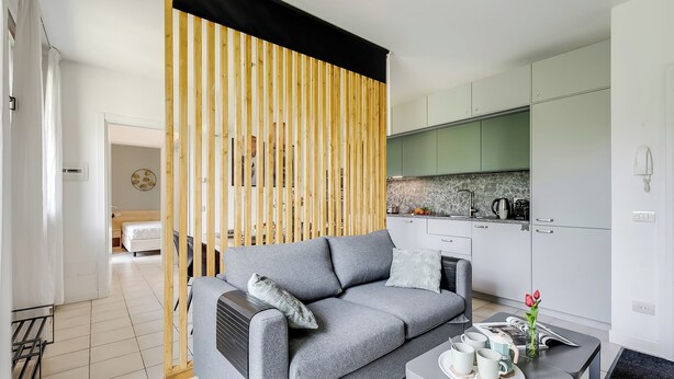 A modern living room with a grey sofa, a wooden partition, a kitchen in the background, and a small table with cups and a vase. - Porto Letizia Roma 4, Porlezza, Italy (Porlezza)