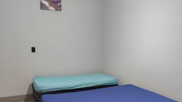 Quarto com duas camas, uma de colchão azul e outra de colchão verde. Há um ventilador de teto e um ponto de luz.