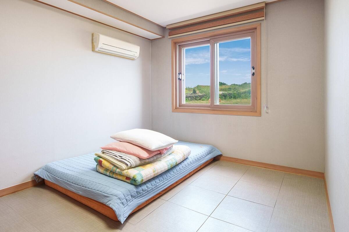 Une petite chambre à coucher, bien rangée, avec un lit simple, une fenêtre donnant sur un espace verdoyant et un climatiseur fixé au mur.