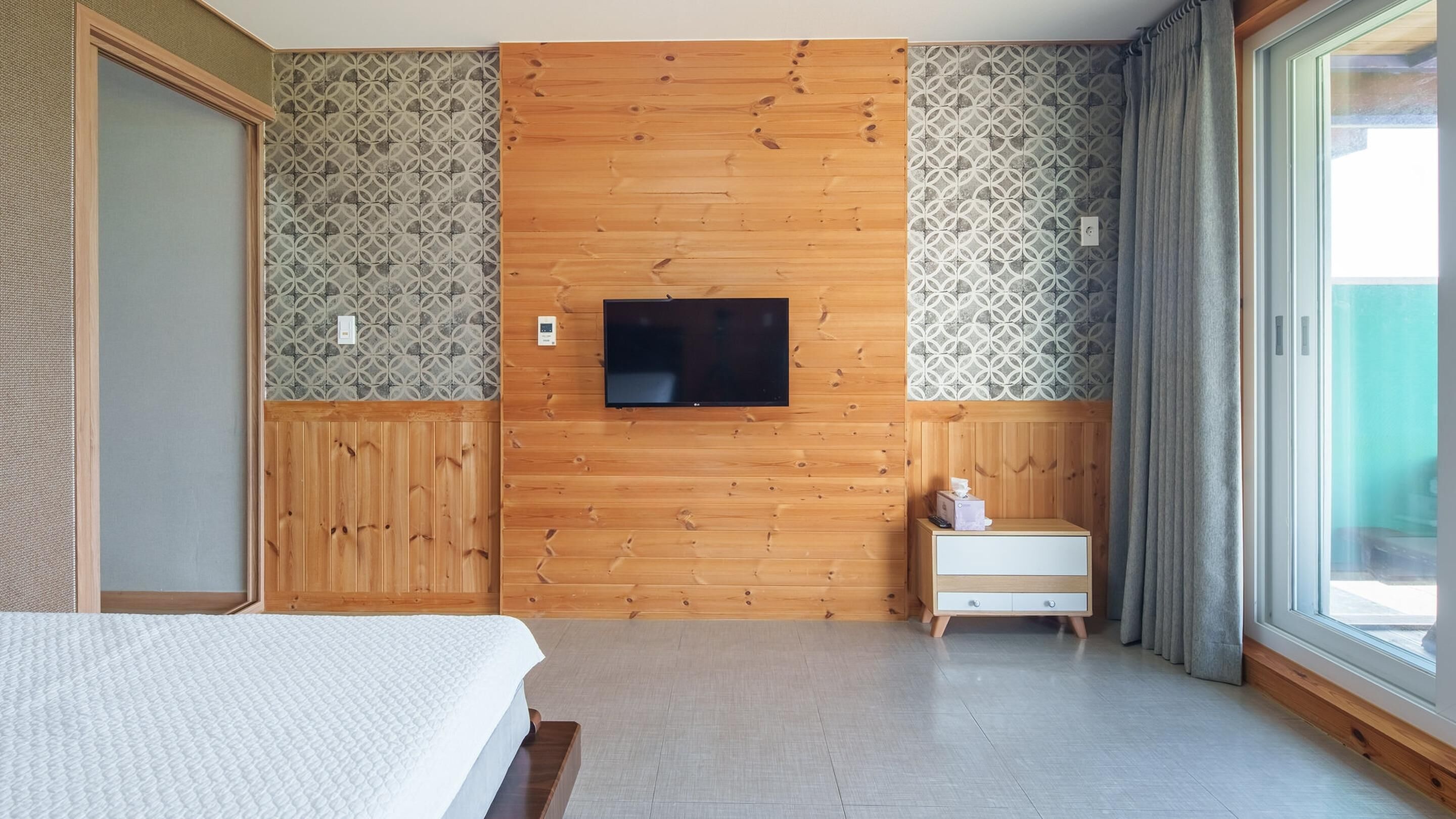 A hotel room with a wooden panel wall, a flat-screen TV mounted above a wooden nightstand, a bed with a white bedspread, and a large window with curtains.