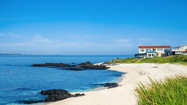 Un hôtel situé sur la côte, avec une plage de sable, un rivage rocheux et une mer d’un bleu limpide.