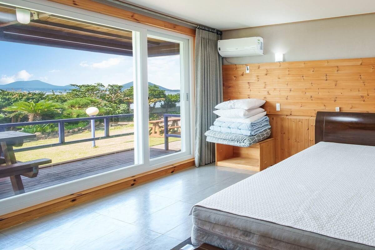 Une chambre avec une grande fenêtre, une tête de lit en bois, un lit avec un matelas blanc et une petite table de chevet avec des coussins.