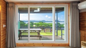 Une pièce dotée d’une porte-fenêtre coulissante donnant sur une terrasse extérieure avec des tables de pique-nique, et offrant une vue sur un lac et des montagnes.