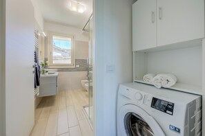 A modern bathroom with a built-in washing machine, a glass shower enclosure, and a view of the outside.