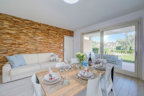 A modern living room with a wooden wall, a white sofa, and a dining area set for four with a view to the outside. - Una Meta Tra Gli Ulivi (Peschiera del Garda)