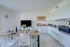 Moderni keittiö, jossa on valkoiset kaapit, puinen ruokapöytä neljälle ja suuri, seinälle asennettu litteänäyttöinen televisio.