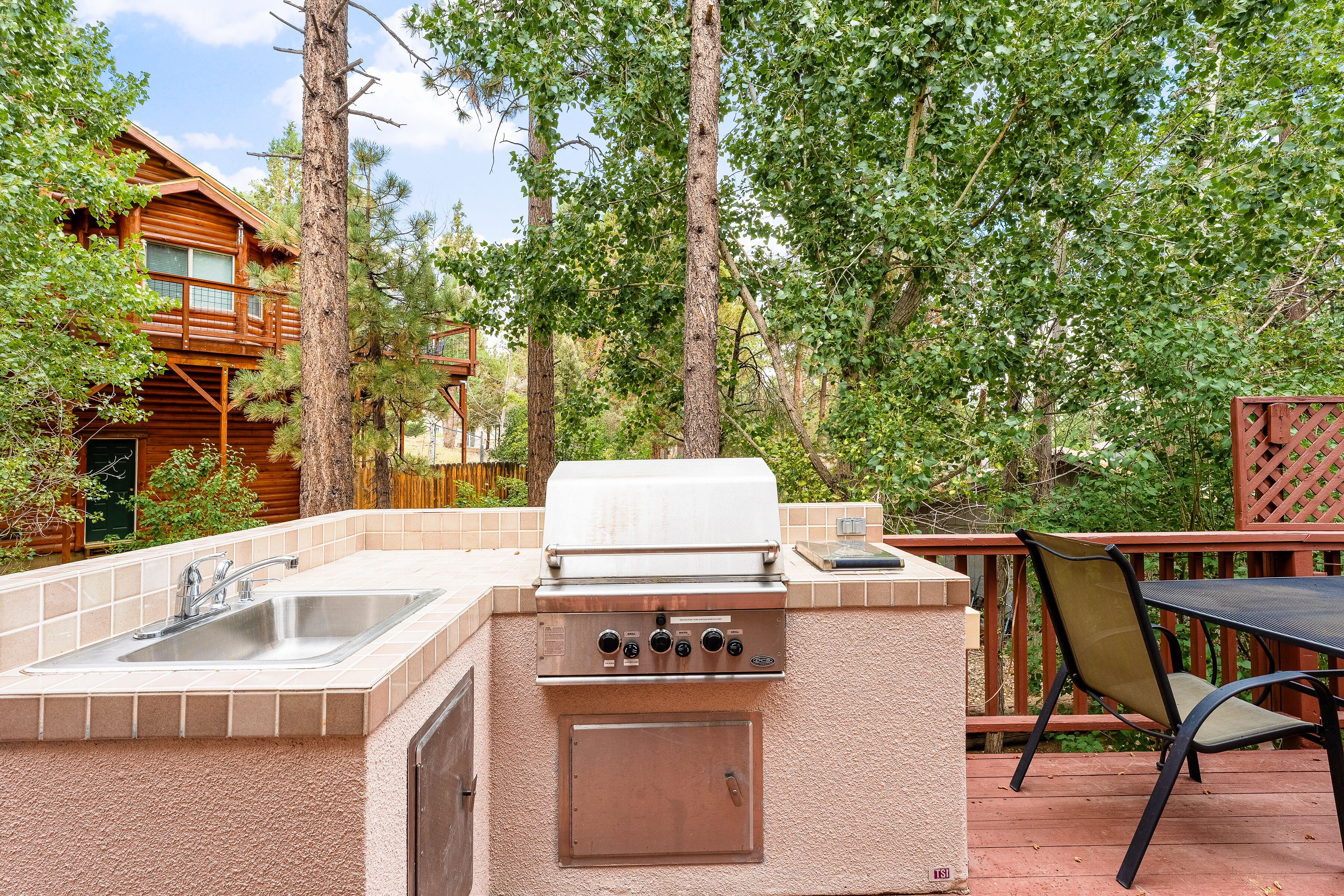 An outdoor kitchen with a sink and grill, surrounded by trees and a wooden deck.