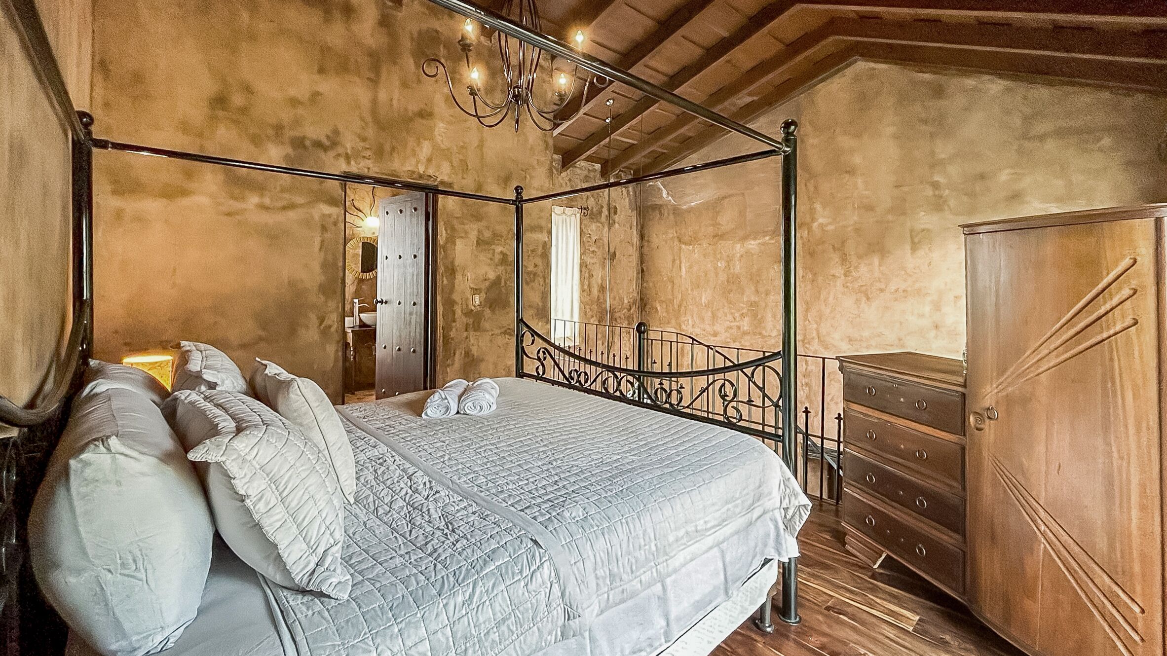 Una cama con dosel, un edredón acolchado y almohadas, un aparador de madera y una araña.