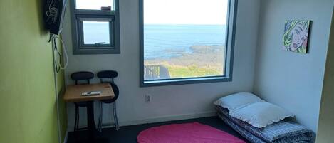 A small room with a bed, a desk, two chairs, a pink mat, and a view of the sea through a large window.
