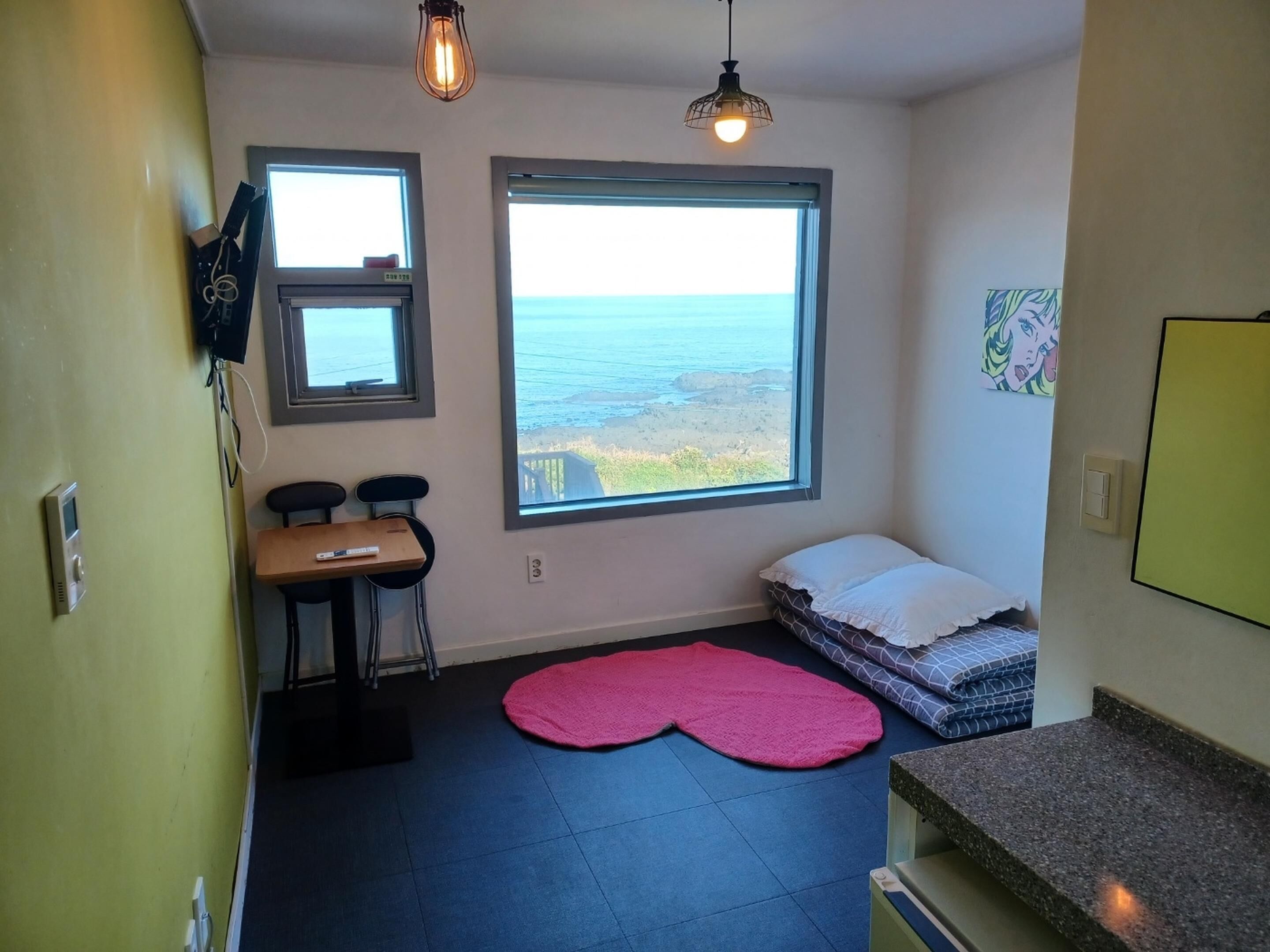 A small, compact room with a large window overlooking the sea, a desk with a chair, a bed with a pink rug, and a kitchenette area.