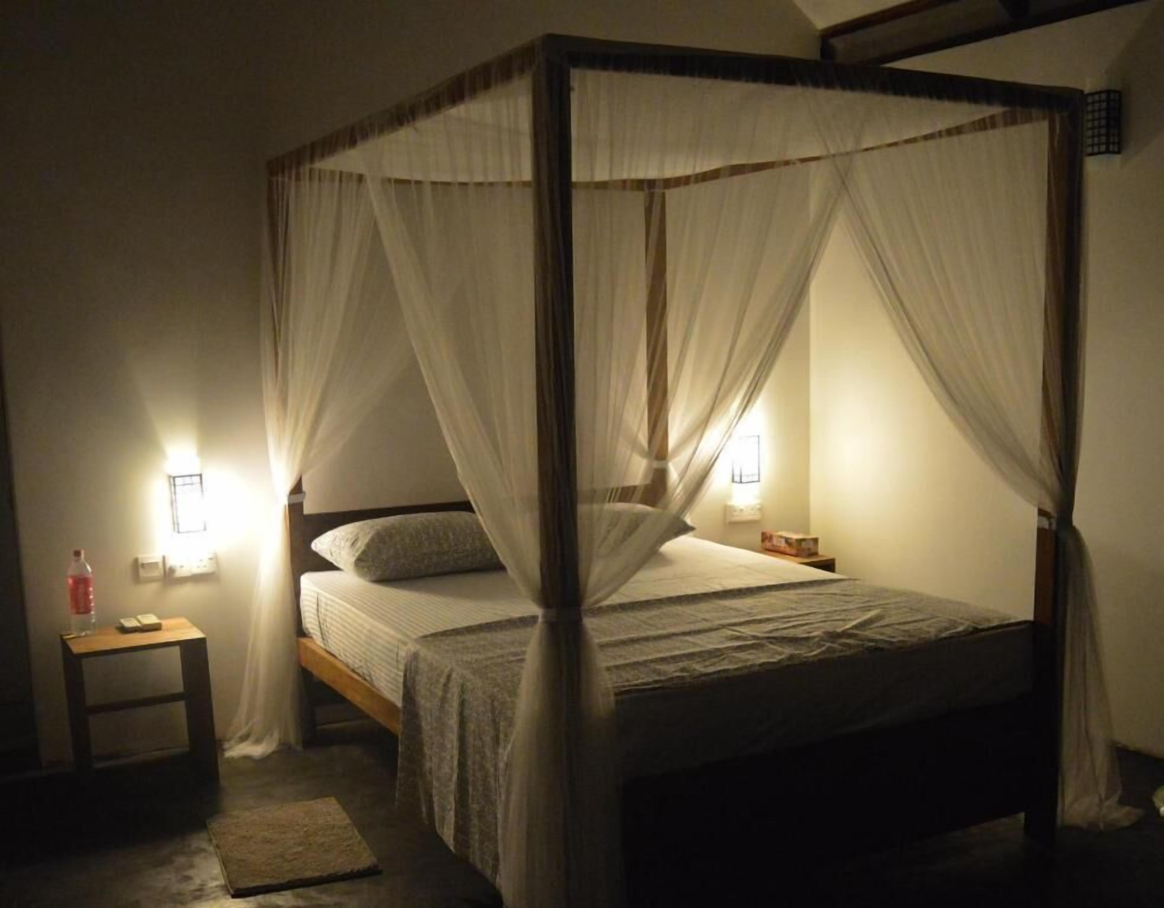A four-poster canopy bed with white bedding, two bedside lamps, and a small wooden nightstand with a bottle and a book.