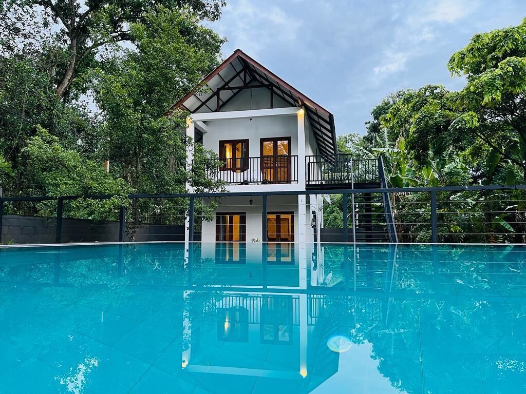 Uma casa de dois andares com varanda sobre uma piscina, rodeada por vegetação exuberante.