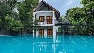 Uma casa de dois andares com varanda sobre uma piscina, rodeada por vegetação exuberante.