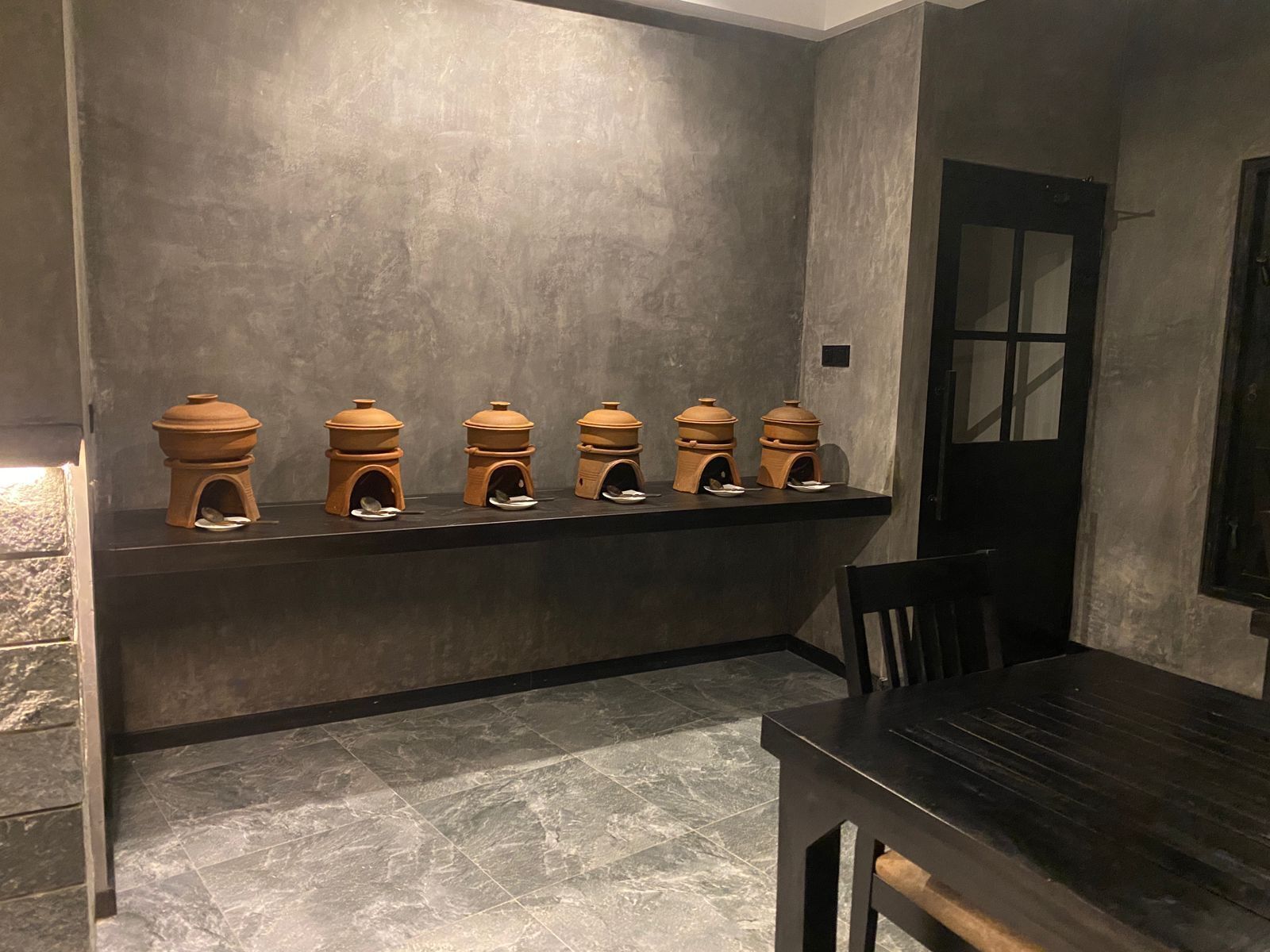 Un interior moderno con una larga mesa de madera oscura y una fila de pequeños objetos en forma de cúpula, de color terracota, sobre una estantería frente a una pared gris con textura.