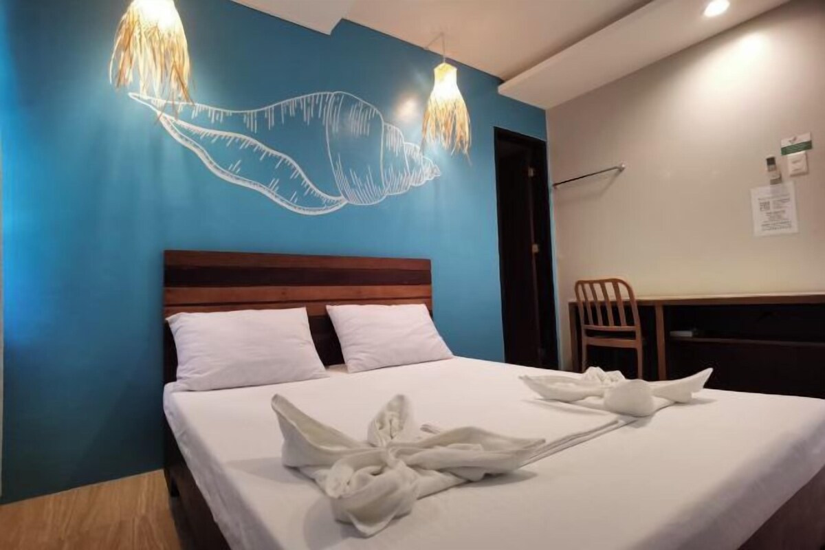 Habitación de hotel con cama, cabecera de madera, pared azul decorativa y lámpara fijada a la pared.