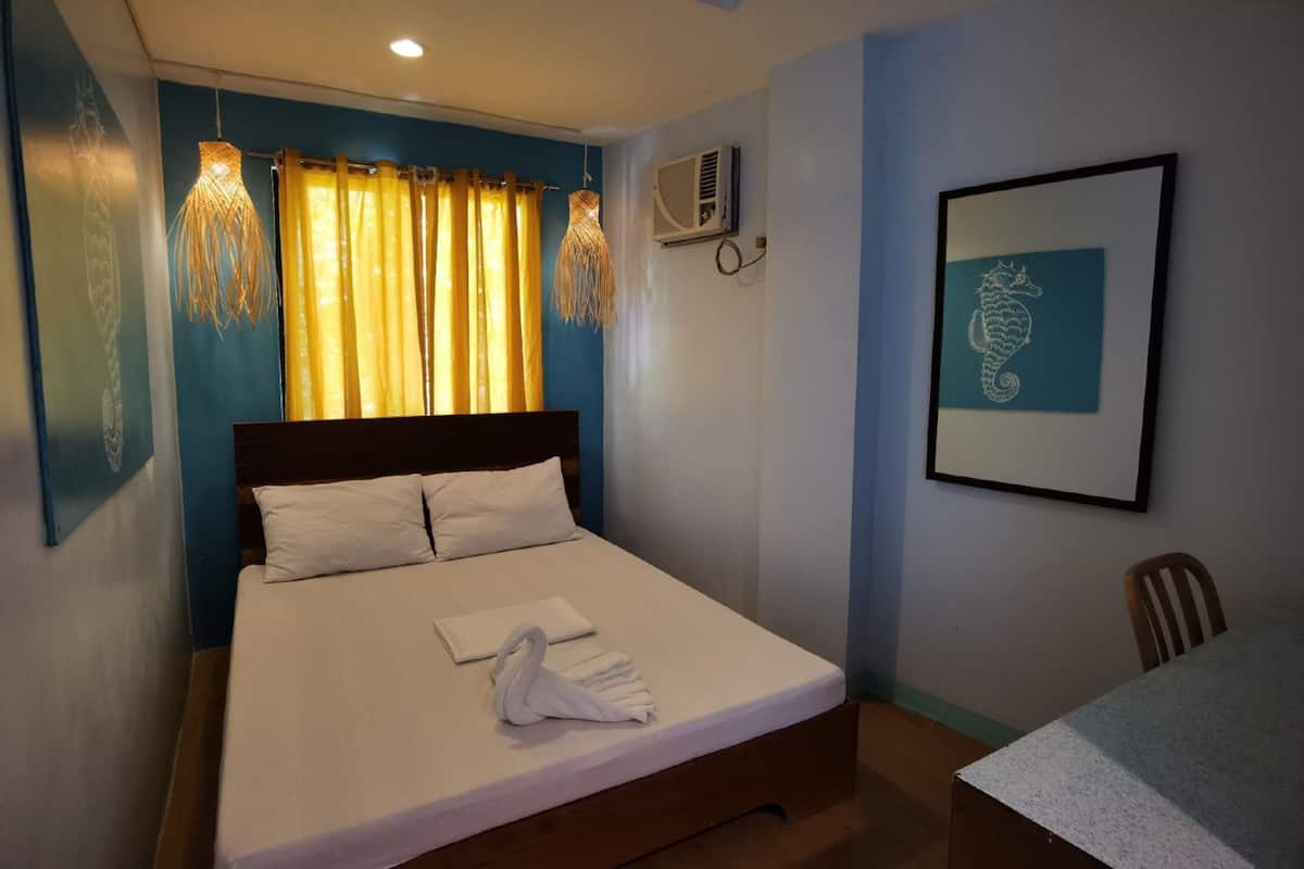 A neatly made bed with white linens and a towel, a wooden headboard, a wall-mounted air conditioner, and a framed seahorse artwork.