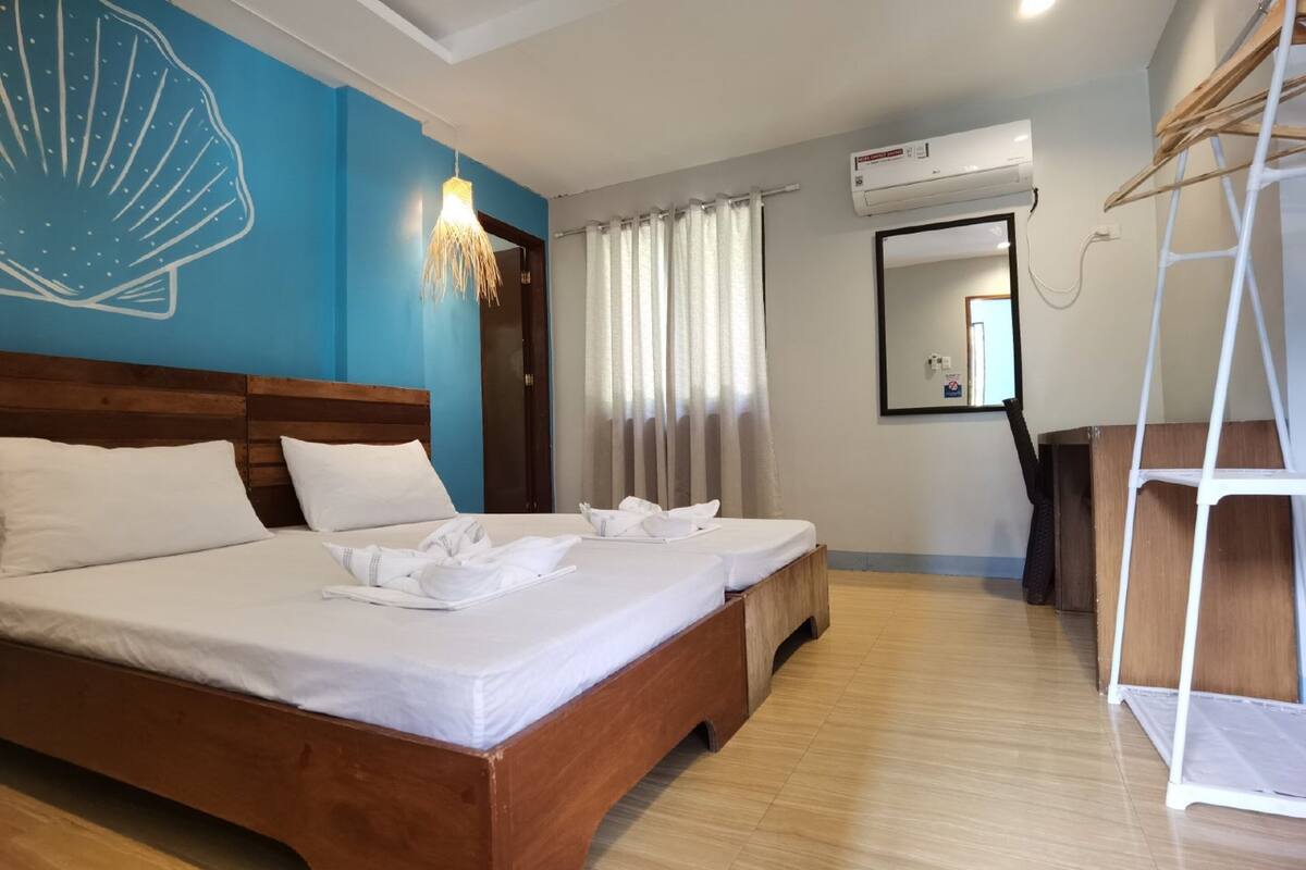 A hotel room with a large bed, a wooden headboard, a blue accent wall, a ceiling fan, a window with curtains, and a white air conditioning unit.