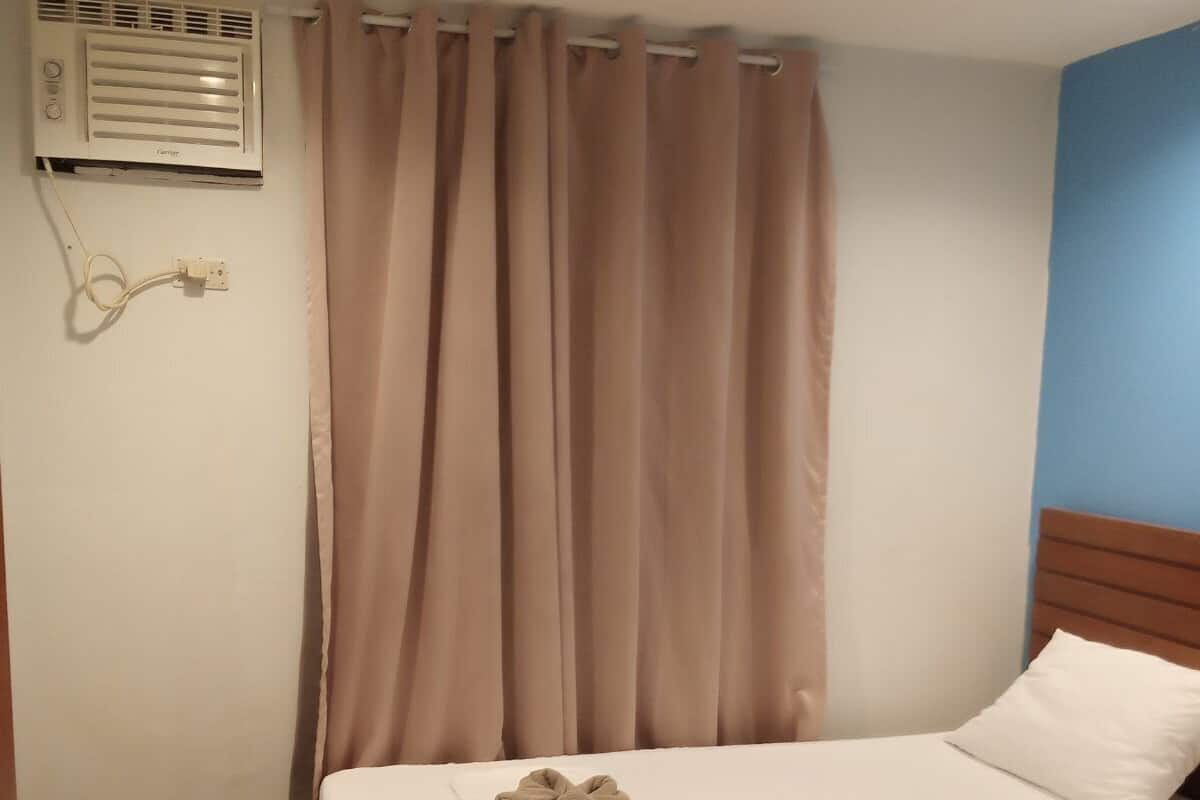A hotel room with two beds, beige curtains, a white air conditioning unit, and a wooden headboard.