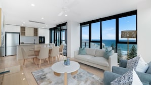 A modern living space with a kitchen, dining area, and a sofa facing a large window with a view of the sea. - Sandbar Burleigh (Burleigh Heads)