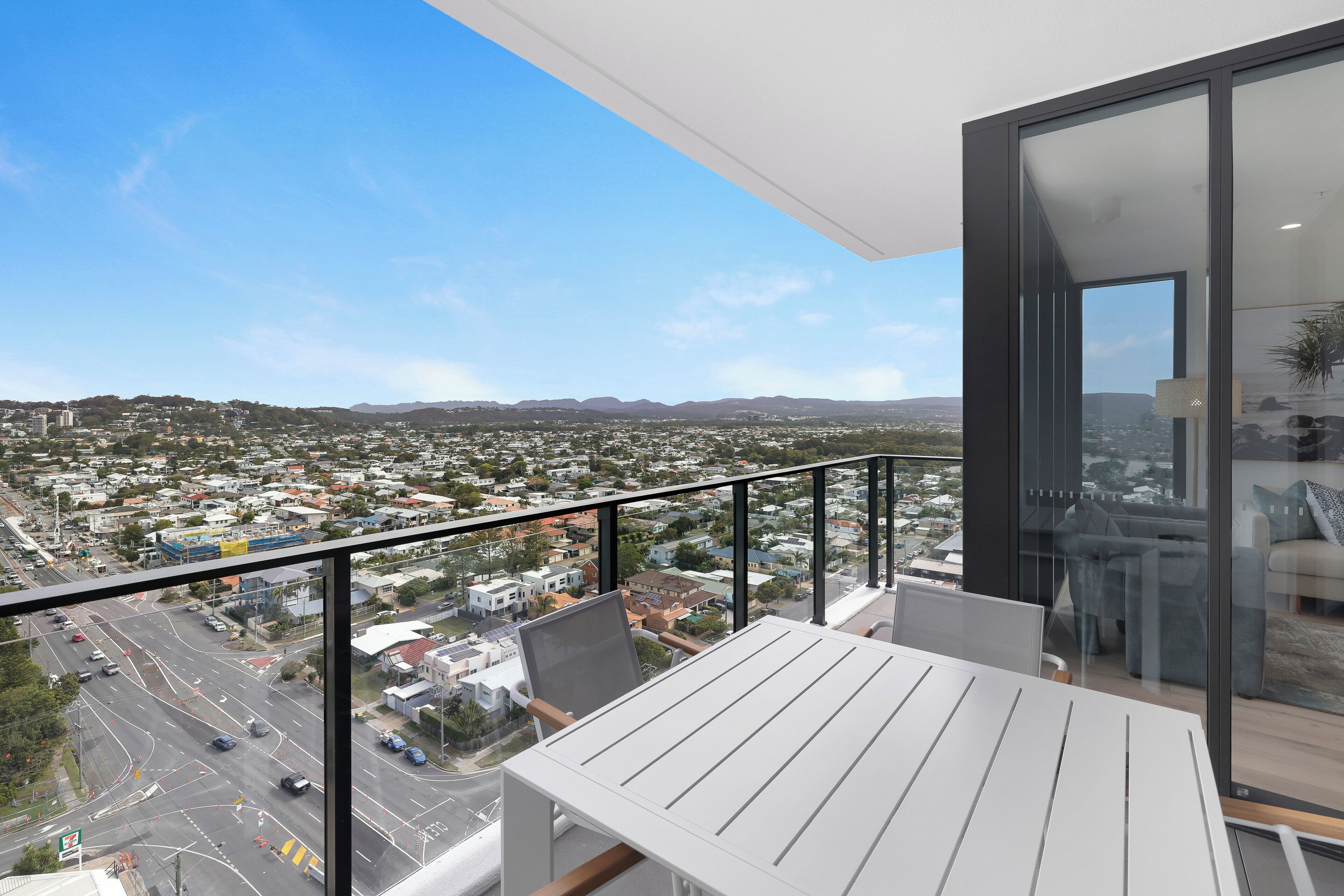 A balcony with a white table and chairs, overlooking a cityscape with buildings and a road.