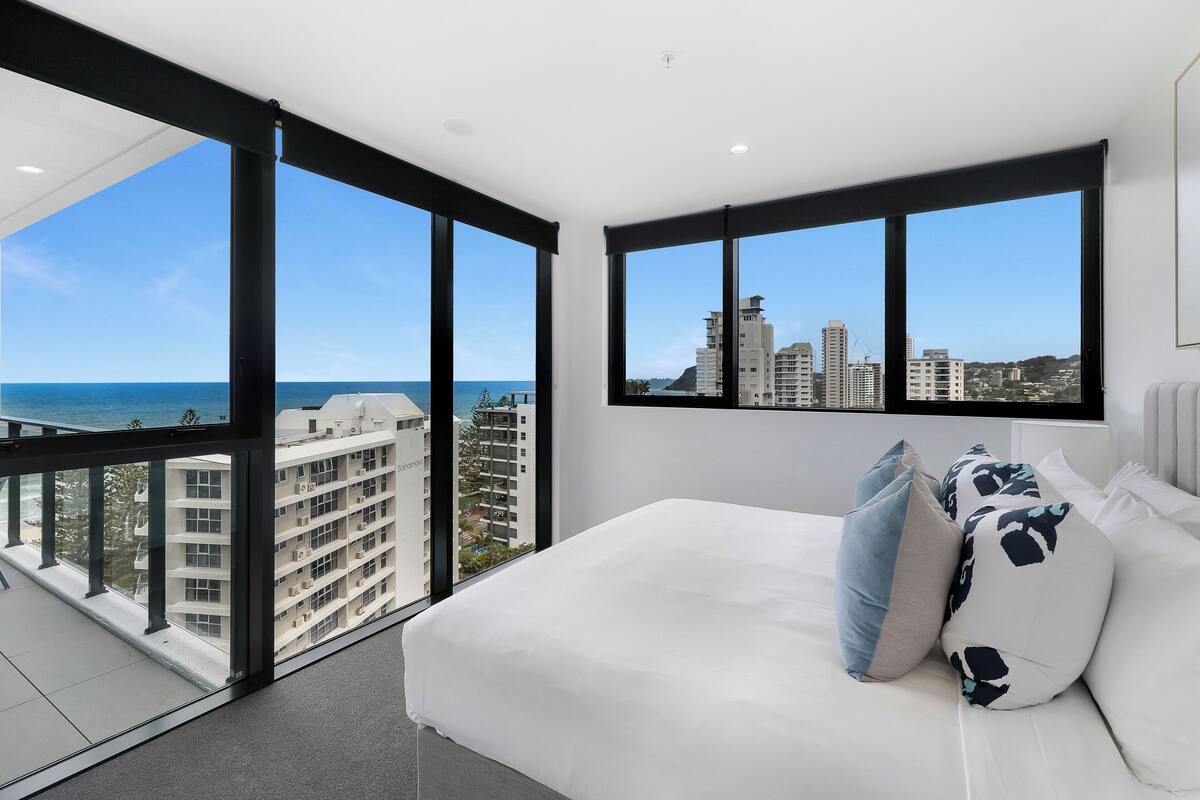 A modern bedroom with a large bed, white bedding, and a view of the city and ocean through floor-to-ceiling windows.