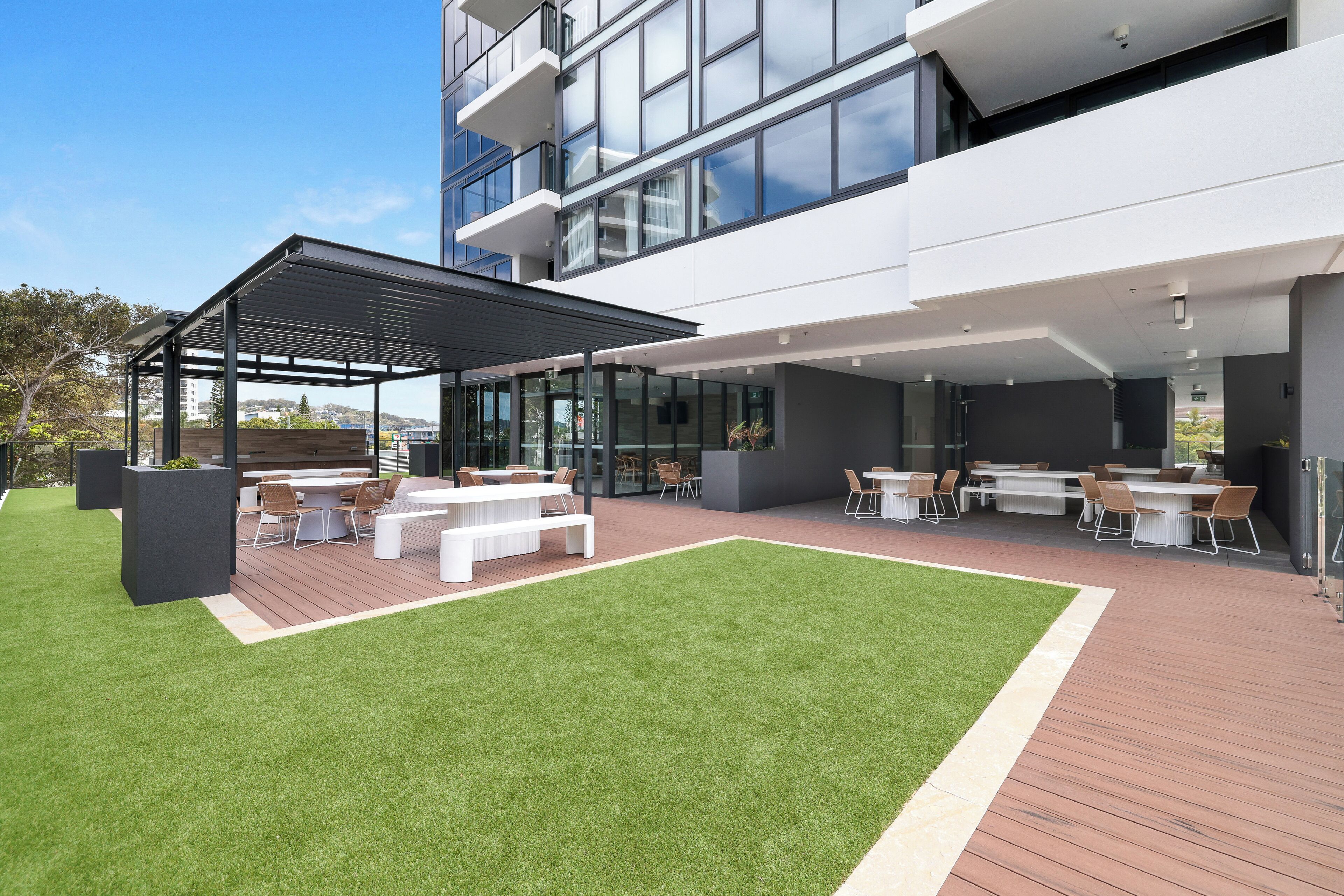 A modern outdoor patio with a covered seating area, a grassy lawn, and a view of a high-rise building.