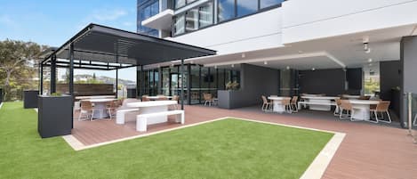 A modern outdoor patio with a covered seating area, a grassy lawn, and a view of a high-rise building.