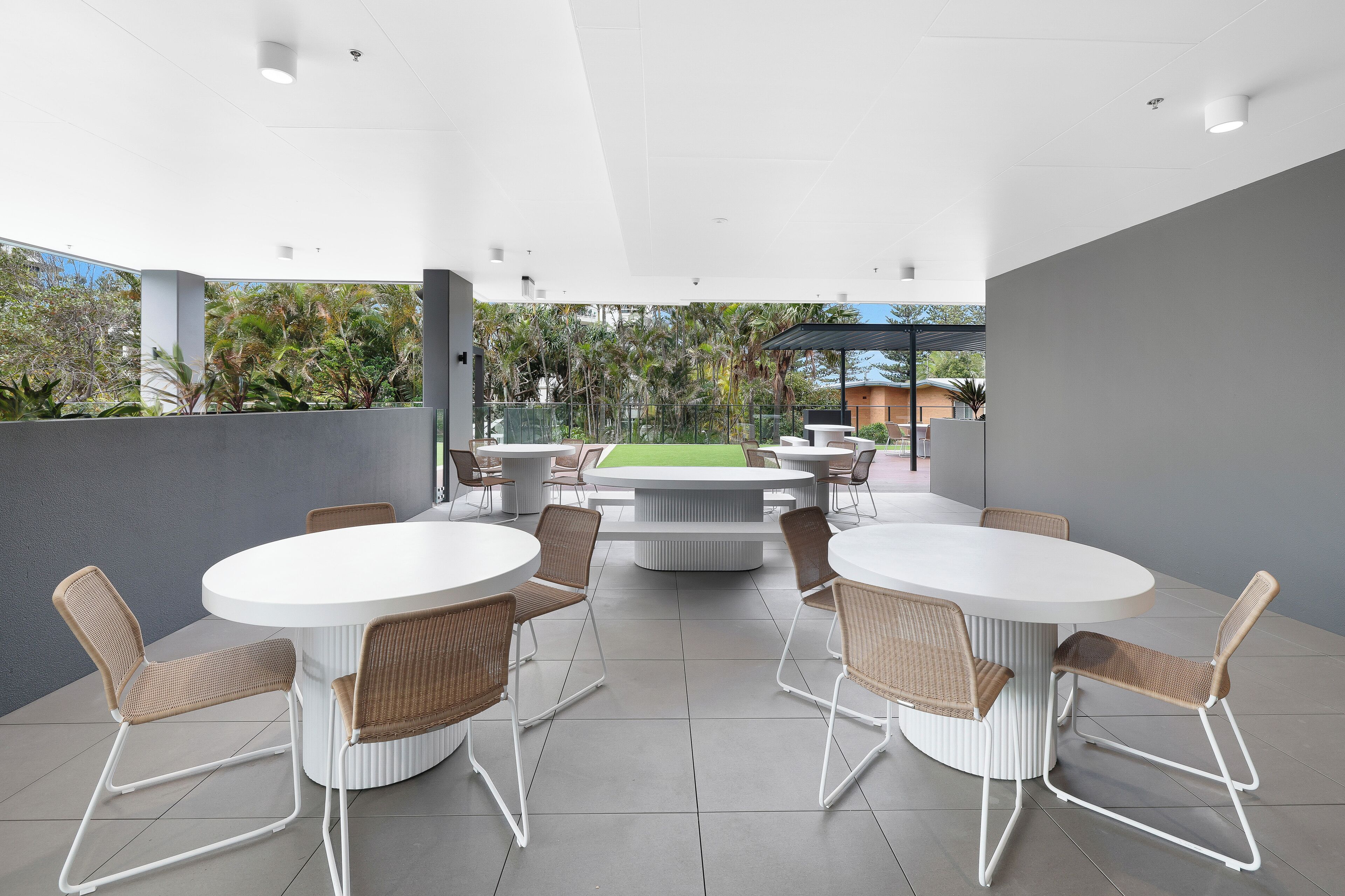 Un espace de restauration extérieur moderne, avec des tables rondes et des chaises en osier, entouré d’un mur gris et offrant une vue sur la verdure.