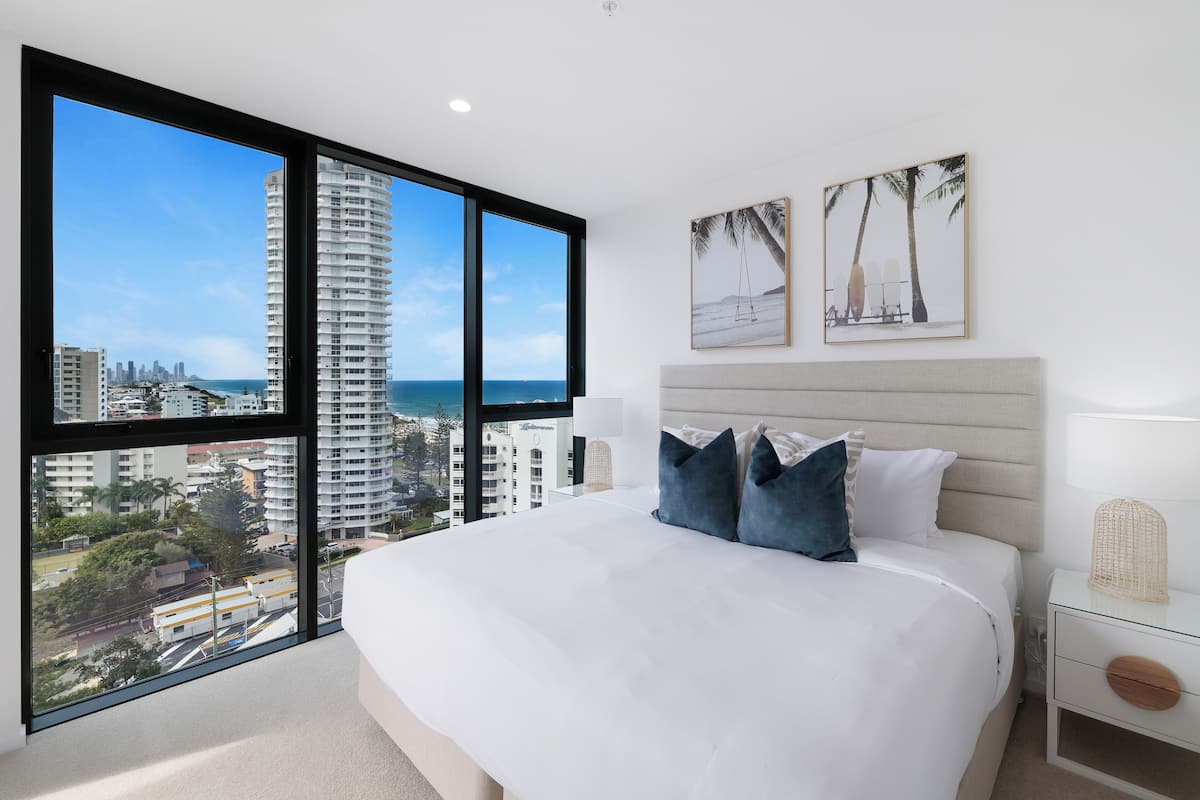 A modern bedroom with a large bed, a nightstand, and a view of the city and ocean.