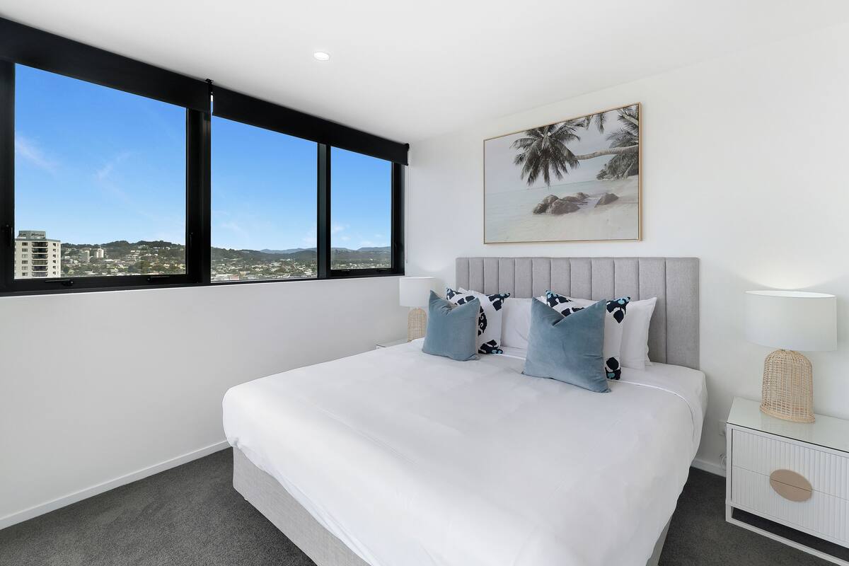 A modern bedroom with a large bed, a view of the city, and a beach scene painting.