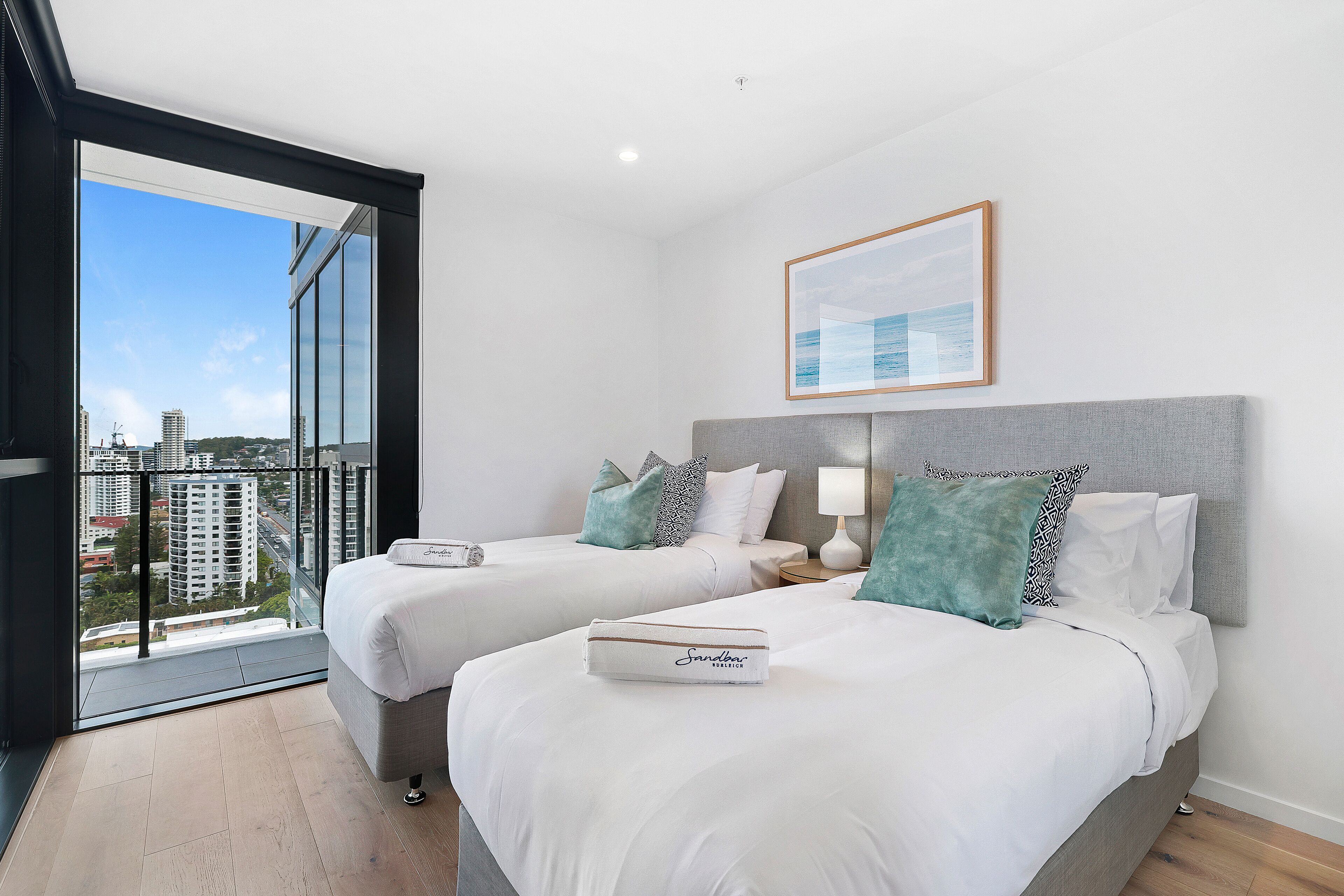 A modern hotel room with two beds, a large window with a city view, and a framed picture on the wall.