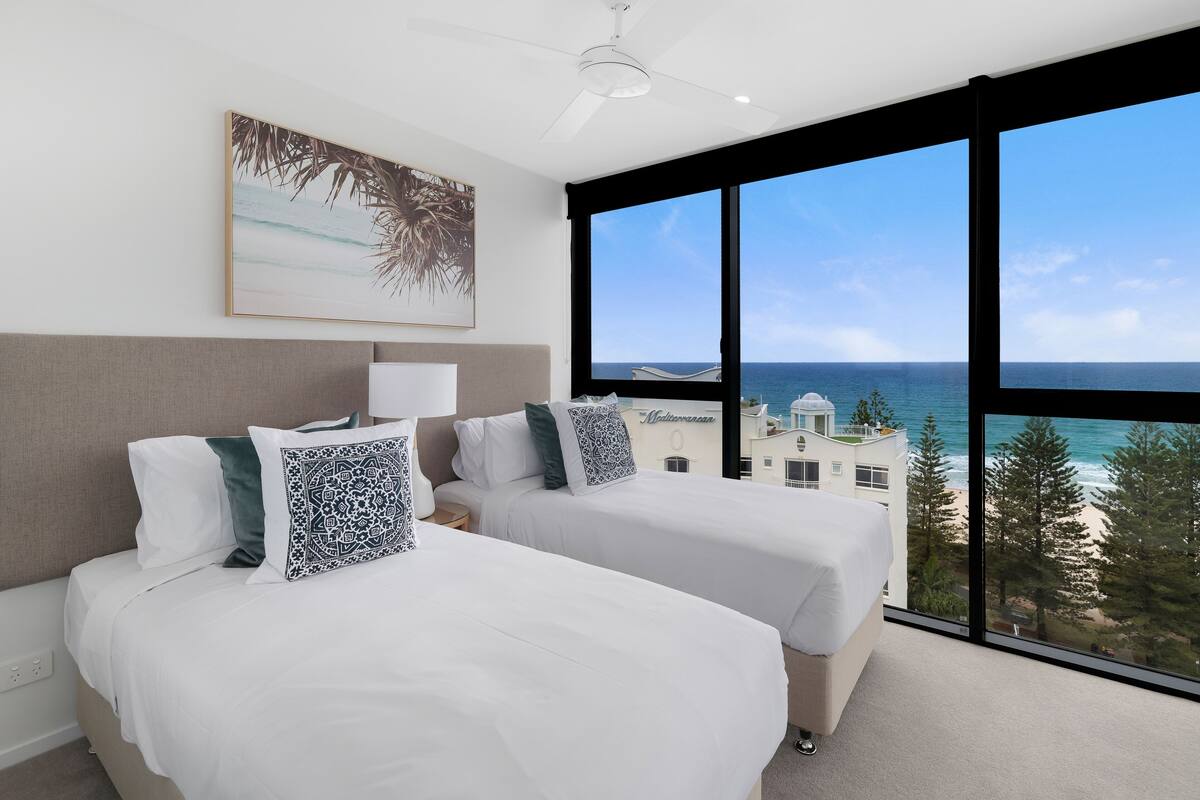 A hotel room with two beds, a ceiling fan, a large window with a view of the ocean, and a painting on the wall.