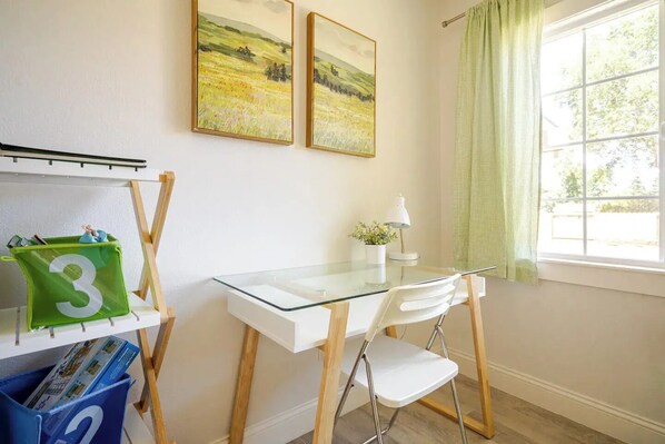 Un estudio con escritorio de vidrio, sillas blancas, una lámpara y dos cuadros de paisajes en la pared.