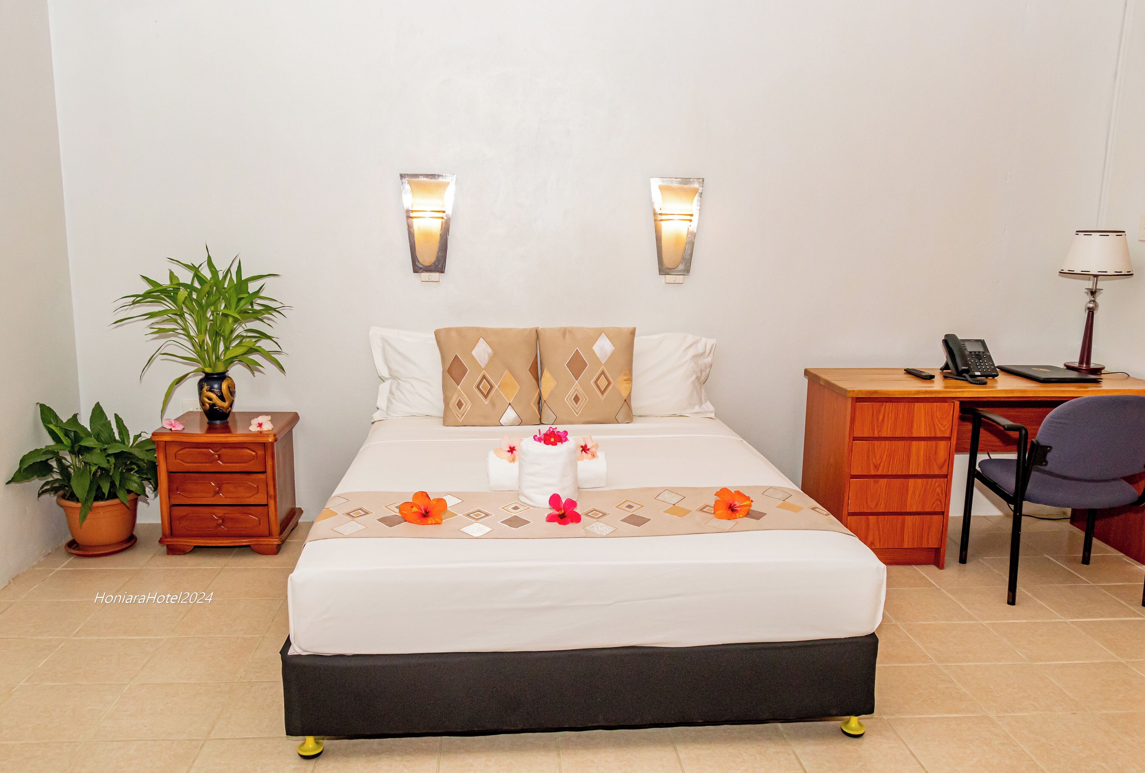 Habitación de hotel ordenada con cama, mesita de noche, escritorio, silla y una planta en maceta.