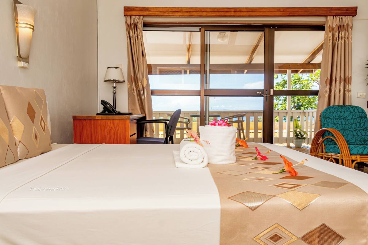 A hotel room with a large bed, a desk with a lamp, a chair, a balcony with a view, and a window with curtains.
