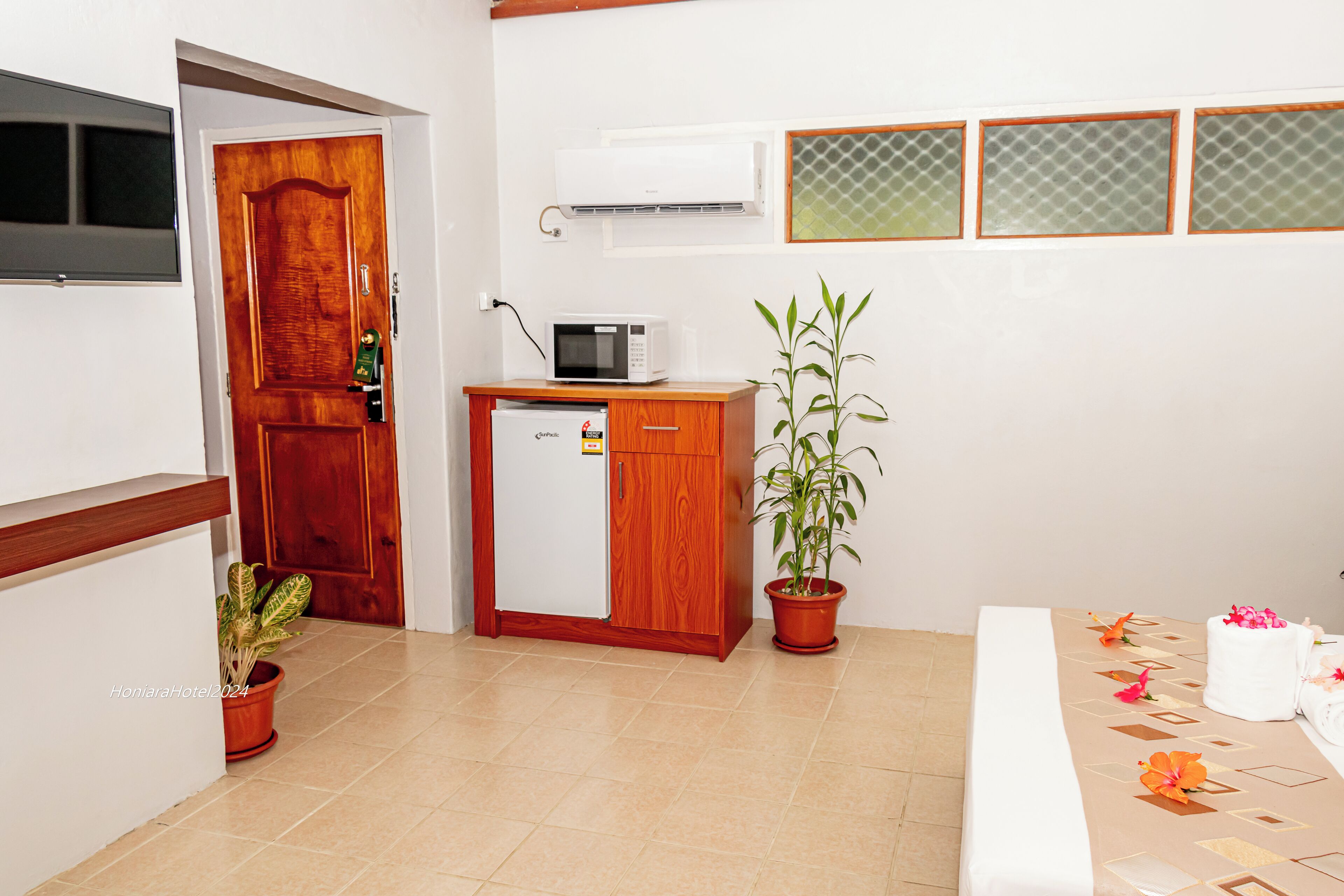 A compact hotel room with a wooden door, a small refrigerator, a microwave, a potted plant, and a bed with a decorative bedspread.