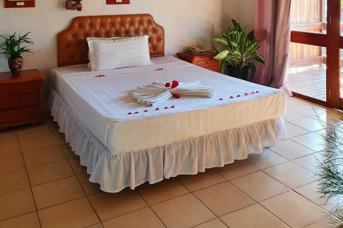A bedroom with a large bed, wooden headboard, two bedside tables, a potted plant, and a sliding glass door leading to an outdoor area.