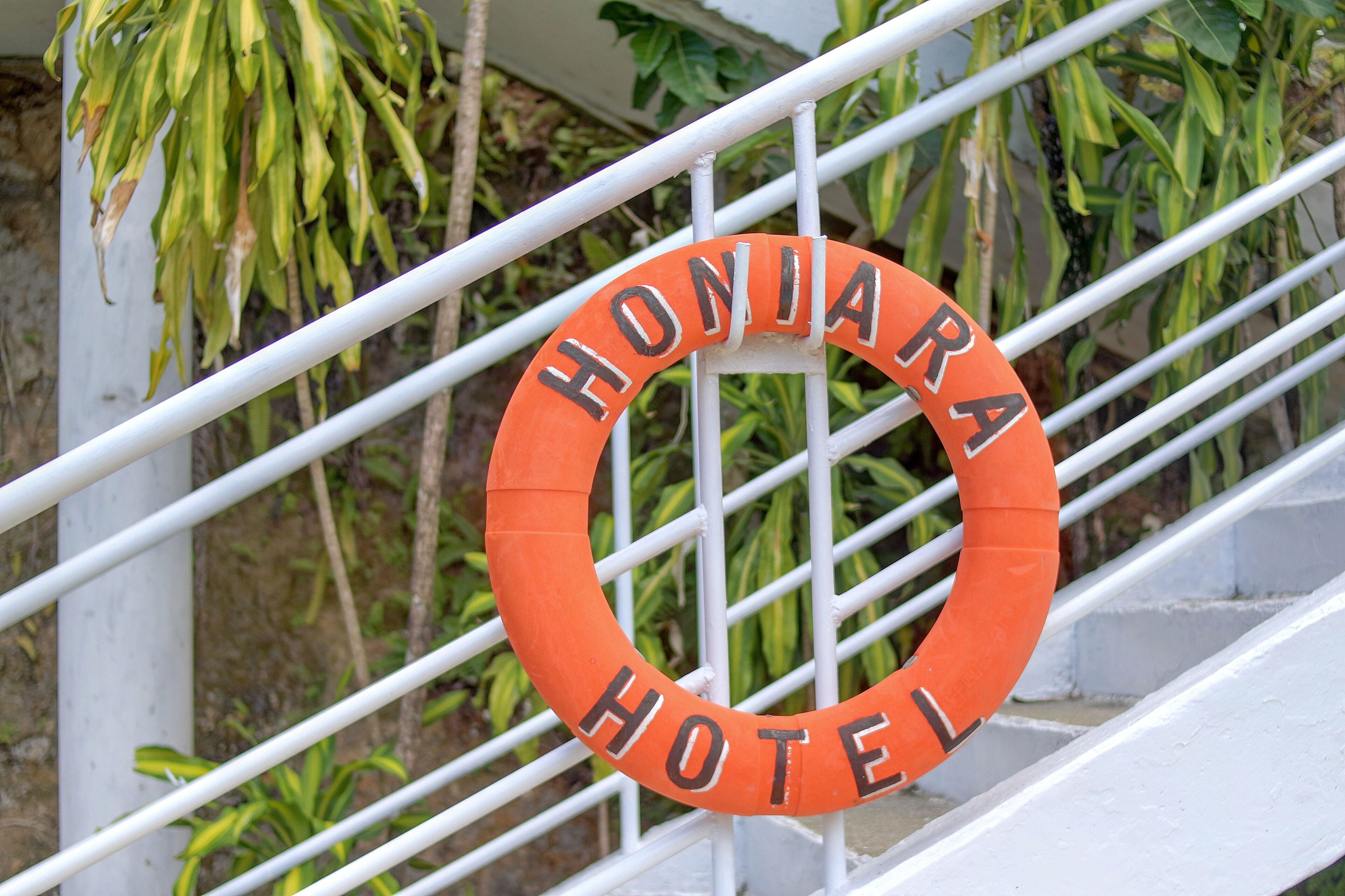 Et orange, rundt skilt med ordene "HONIARA HOTEL" på gelænderet af en hvid metal trappe.