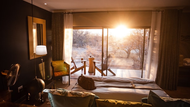 A hotel room with a bed, a chair, a table, and a view of trees through a large window.