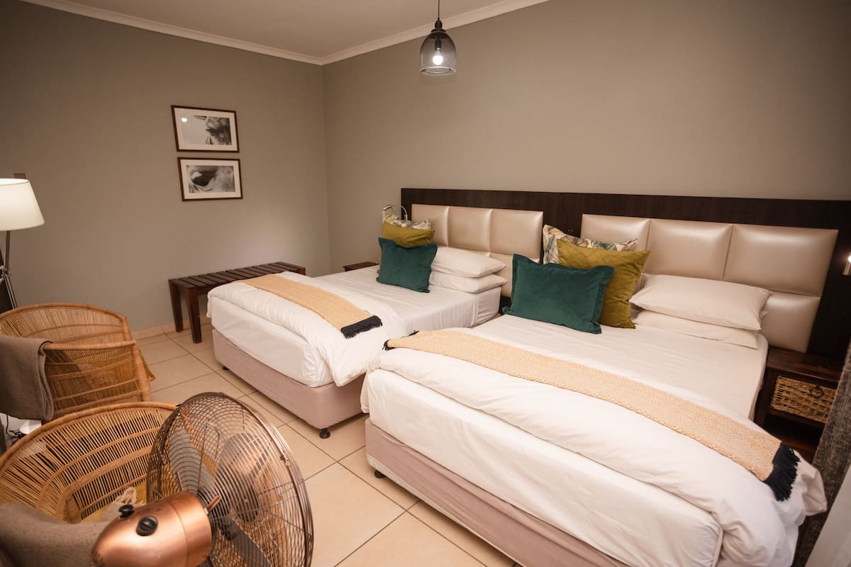 Quarto de hotel com duas camas, área de estar com uma cadeira, luminária e ventilador.