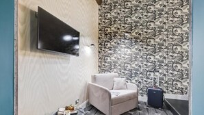 A room with a flat-screen TV mounted on the wall, a patterned wallpaper, a grey sofa, a small ottoman, and a suitcase. - Premium Private Suites (Rome)