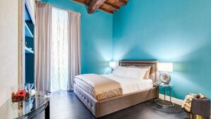A bedroom with a bed, a glass table with a flower vase, a round side table with a lamp, and a window with curtains. - Premium Private Suites (Rome)