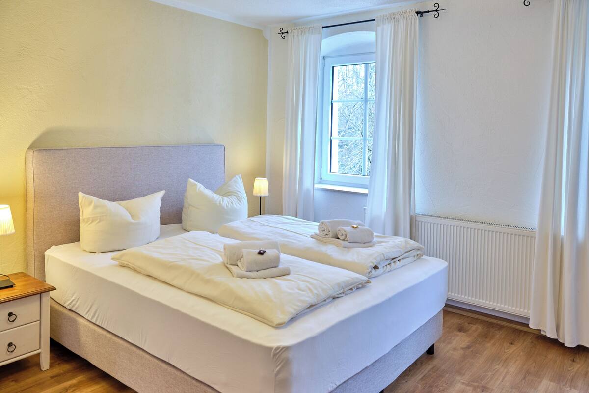 Una cama doble con sábanas blancas, una mesita de noche con lámpara y una ventana con cortinas.