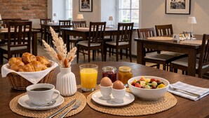 Un desayuno con croissants, café, jugo, huevos y un bol de fruta sobre una mesa en un comedor con sillas y mesas de madera, paredes de ladrillo y cuadros en las paredes.