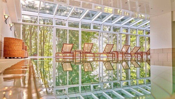Un amplio espacio interior con techo de vidrio que deja entrar luz natural, y una fila de sillas de madera frente a un piso reflectante.