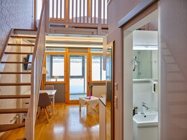 Un interior moderno con una escalera de madera, una pequeña mesa con un florero y un baño con lavamanos y espejo.