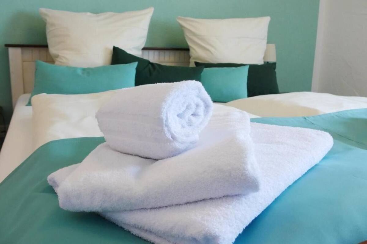 Una cama bien hecha con ropa de cama blanca y aguamarina, una toalla blanca enrollada y almohadas a juego.