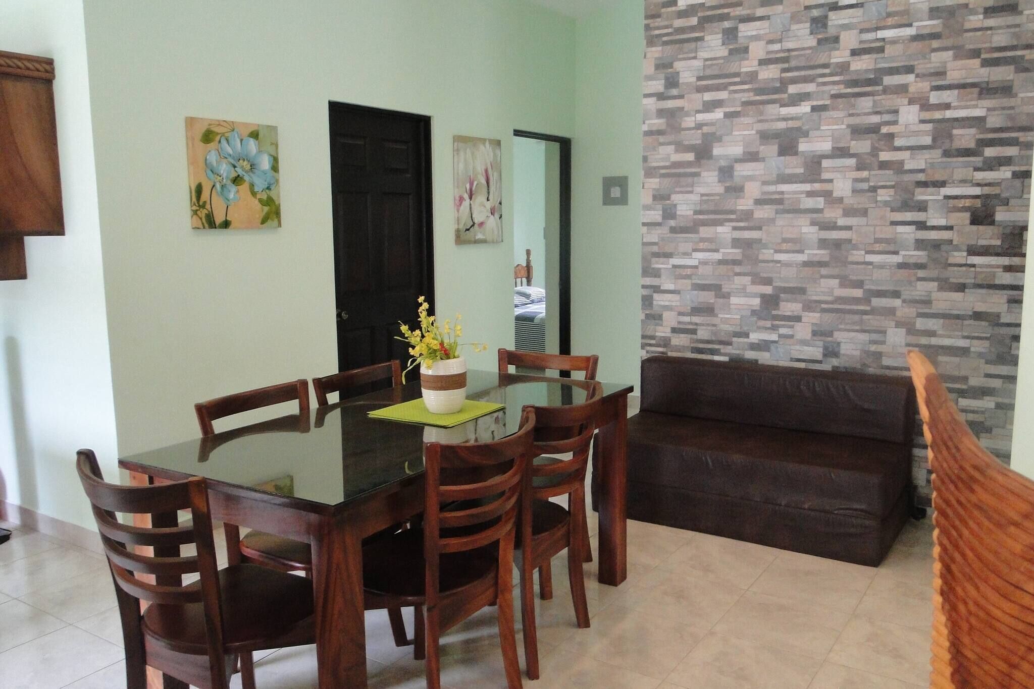 A dining area with a glass table, wooden chairs, a brown sofa, and a decorative vase with flowers.